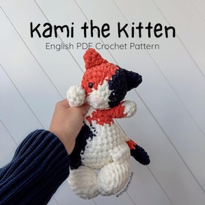 Peut inclure: Un jouet de chaton crocheté à la main, blanc, rouge et bleu marine. Le chaton a un nez rose. Le texte "Kami the Kitten" et "English PDF Crochet Pattern" sont affichés au-dessus du chaton. Le jouet est tenu par une personne.