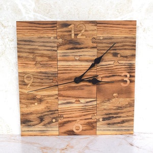 May include: A wooden clock with black hands and Roman numeral markings on a brown wood background. The clock is made of puzzle pieces.