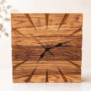 May include: A square wooden wall clock featuring a natural light brown wood grain finish. The clock face has deep, carved radial grooves extending from the center toward the edges. Three thin black metal hands are positioned in the center.