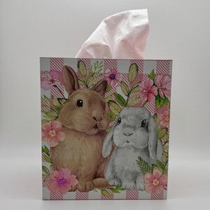 May include: A decorative tissue box featuring a brown and gray rabbit design with pink flowers and green leaves. The box has a pink and white checkered pattern and a stack of pink tissues.