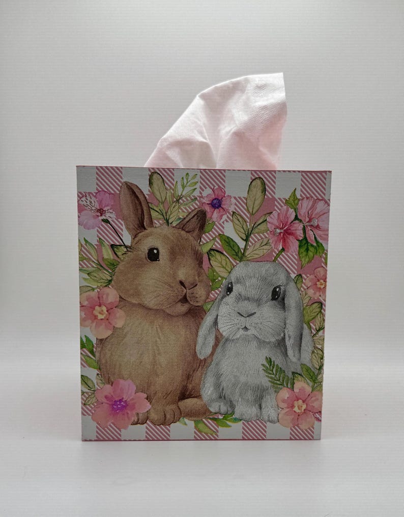 May include: A decorative tissue box featuring a brown and gray rabbit design with pink flowers and green leaves. The box has a pink and white checkered pattern and a stack of pink tissues.