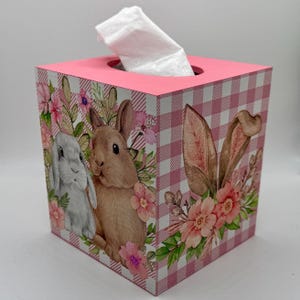 May include: A square tissue box with a pink top and a pink and white gingham pattern. The sides feature illustrations of bunnies, flowers, and greenery. A white tissue is pulled from the top opening.