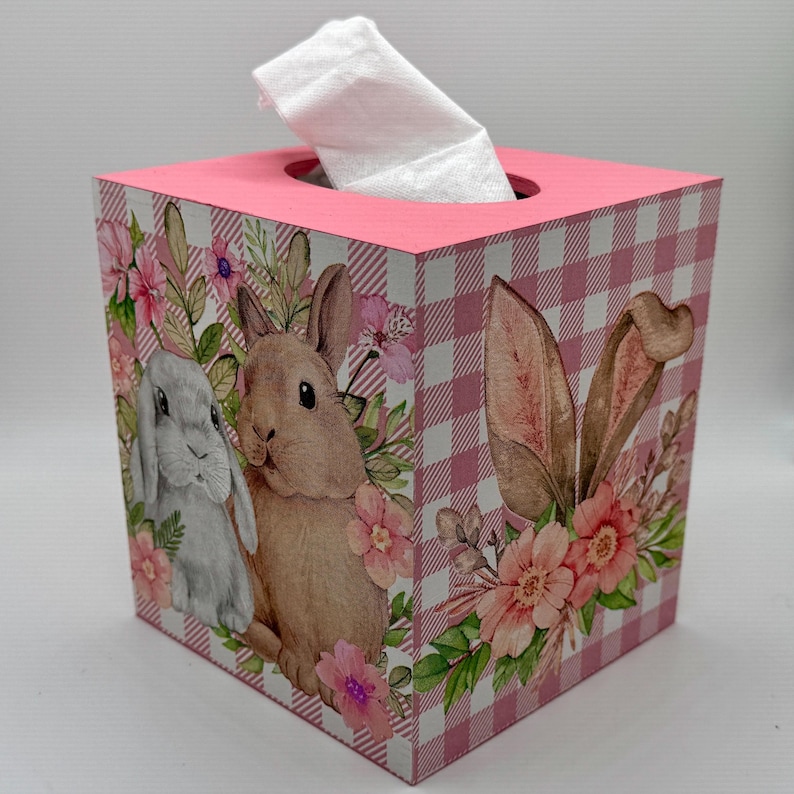 May include: A square tissue box with a pink top and a pink and white gingham pattern. The sides feature illustrations of bunnies, flowers, and greenery. A white tissue is pulled from the top opening.
