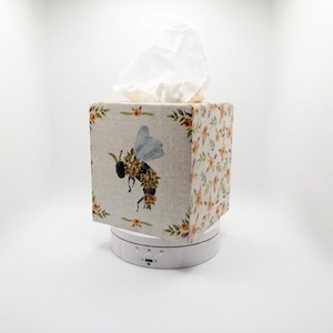May include: A square tissue box cover with a bee illustration. The box has a light beige background with a floral pattern and a bee design on one side. The bee has blue wings and is surrounded by yellow and orange flowers. White tissues are visible.