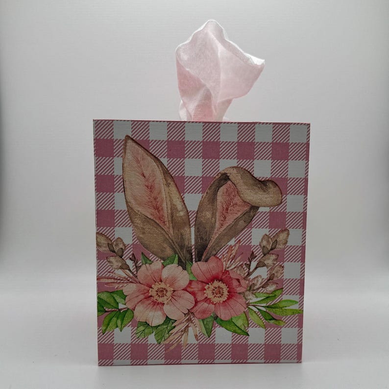 May include: A decorative tissue box featuring a pink and white gingham pattern. The box is adorned with a floral arrangement and bunny ears. Pink tissue paper is pulled from the top, adding a touch of color. A charming Easter-themed accent.