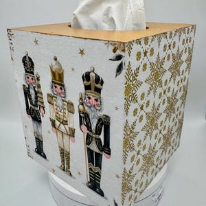 May include: A square tissue box featuring a festive design. The box is white with gold accents and illustrations of nutcracker soldiers. The right side of the box has a gold snowflake pattern. Tissues are visible at the top.