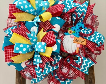 May include: A red, white, and blue wreath with a gnome holding a sign that says "Hello!" The wreath is made of mesh and has a variety of ribbons, including polka dot, chevron, and plaid.