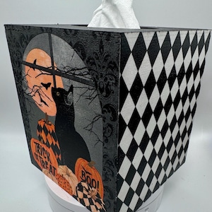 May include: A decorative tissue box with a Halloween theme, featuring a black cat, pumpkins, and the phrases "Trick or Treat" and "Boo!". One side of the box has a black and white diamond pattern.