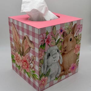 May include: A square tissue box with a pink gingham pattern and a pink top. The sides feature illustrations of bunnies and flowers. White tissues are visible at the top.
