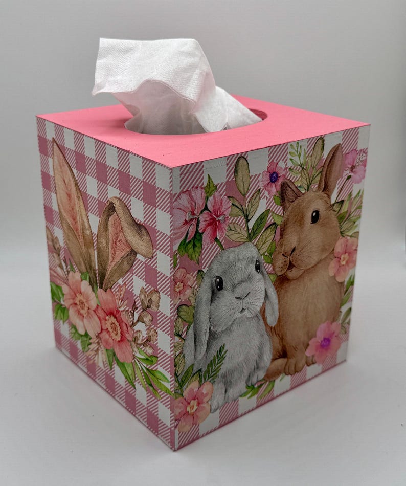 May include: A square tissue box with a pink gingham pattern and a pink top. The sides feature illustrations of bunnies and flowers. White tissues are visible at the top.