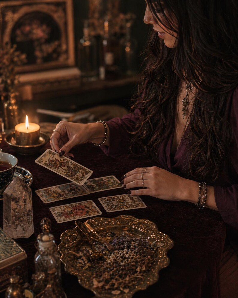 May include: A set of illustrated tarot cards with intricate designs, featuring one card being held above a dark velvet surface. The cards display detailed artwork in muted tones, accompanied by a lit candle, a clear crystal, and a gold-toned tray.