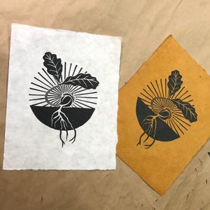 May include: Two linocut prints of a sprouting seed design. One print is on white paper, the other on mustard-colored paper. The black and white design features a seed with roots, leaves, and a sunburst pattern.