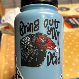 May include: A light blue water bottle with a black lid and handle. The bottle features a sticker with a vulture illustration and the text "Bring out your Dead" in black lettering. Additional stickers are visible.