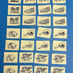 May include: A collection of small, rectangular fabric patches with black linocut prints. The designs include mushrooms, berries, foliage, and animal skulls. The patches are arranged on a blue grid surface.