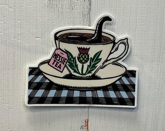 May include: A white teacup with a black Nessie monster teabag, a pink tag that says "Nessie Tea", and a green thistle design. The teacup is on a white saucer with a black and white checkered pattern.