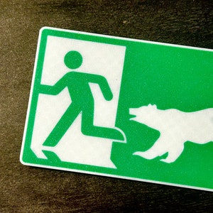 May include: Green and white exit sign with a stick figure running to the left and a white silhouette of a bear running to the right. The sign has a textured surface.