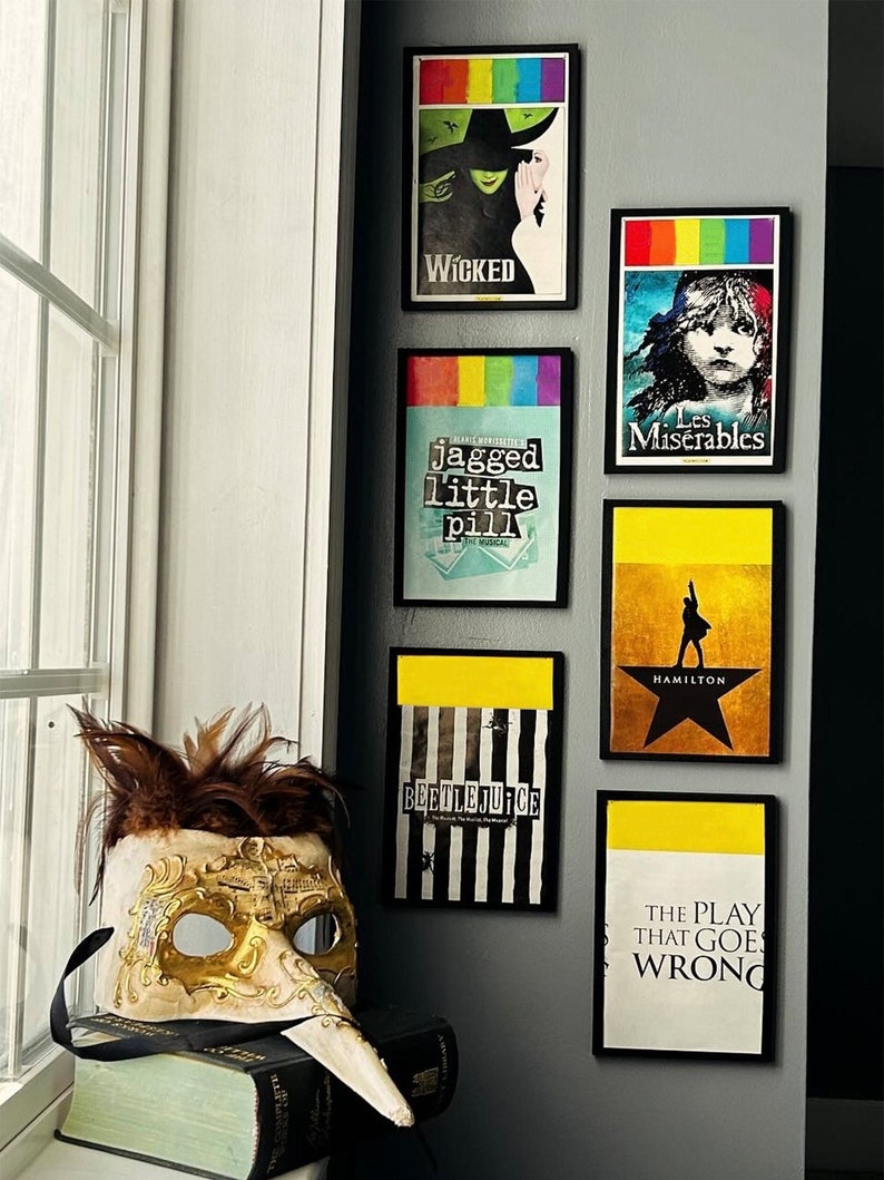 May include: A wall display of framed Broadway show posters, including Wicked, Les Miserables, Jagged Little Pill, Hamilton, Beetlejuice, and The Play That Goes Wrong. A gold and white masquerade mask sits on a book in the foreground.