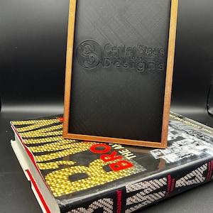May include: A wooden frame with a black background and the text "Center Stage Designs" in white. The frame is sitting on top of a book with the title "Broadway The American Musical".