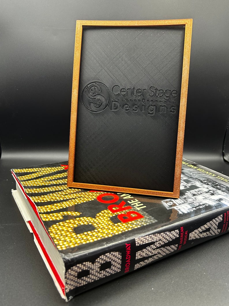 May include: A wooden frame with a black background and the text "Center Stage Designs" in white. The frame is sitting on top of a book with the title "Broadway The American Musical".