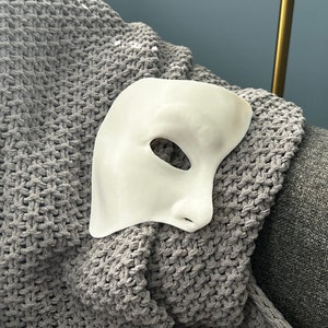 May include: A white plastic masquerade mask with a single eye hole, resting on a grey knitted blanket.