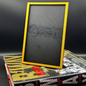 May include: A black picture frame with a gold border. The frame is made of a textured material and has the words "Center Stage Designs" embossed on the front.
