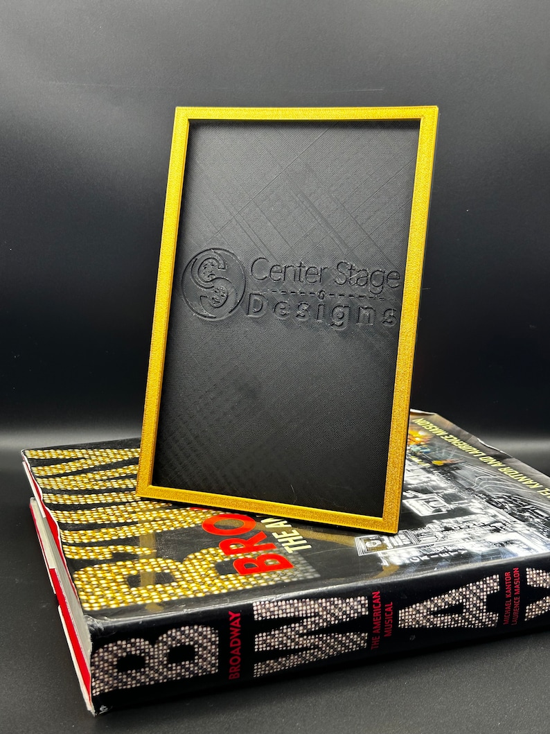 May include: A black picture frame with a gold border. The frame is made of a textured material and has the words "Center Stage Designs" embossed on the front.