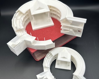 May include: A white 3D printed model of a circular building with multiple levels and a small roofed structure in the center. The model is sitting on a red book.