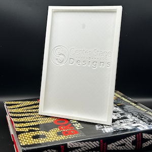 May include: White rectangular frame with a raised, embossed logo that reads "Center Stage Designs". The frame is sitting on top of a book with the word "Broadway" printed on the spine.