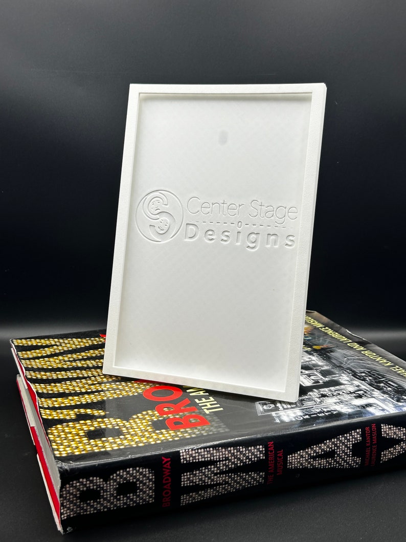 May include: White rectangular frame with a raised, embossed logo that reads "Center Stage Designs". The frame is sitting on top of a book with the word "Broadway" printed on the spine.