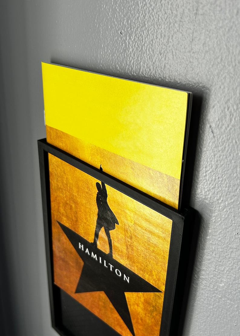 May include: A black framed print of a silhouette of a person standing on a gold star with the word "HAMILTON" written on it. A yellow piece of paper is partially visible behind the frame.