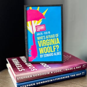 May include: A framed poster advertising a play called "Who's Afraid of Virginia Woolf?" by Edward Albee. The poster is blue with a pink umbrella and a gun. The poster is on top of three books, all by Stephen Sondheim.