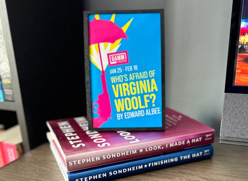 May include: A framed poster advertising a play called "Who's Afraid of Virginia Woolf?" by Edward Albee. The poster is blue with a pink umbrella and a gun. The poster is on top of three books, all by Stephen Sondheim.