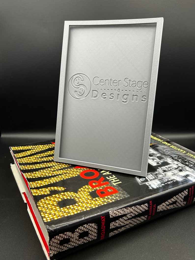 May include: A silver-colored frame with the text "Center Stage Designs" embossed on the front. The frame is sitting on top of a book with the title "Broadway: The American Musical".
