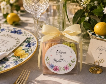 May include: A clear square favor box containing two macarons, tied with a pale yellow ribbon. A circular sticker on the front features a floral wreath, two small pink hearts, and the text: Thank you for sharing our day, Olivia & Matthew.