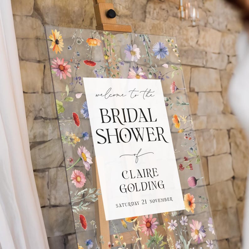 May include: A clear acrylic sign featuring a colorful wildflower print around the border. The center white panel displays the text: welcome to the BRIDAL SHOWER of CLAIRE GOLDING SATURDAY 21 NOVEMBER. The sign is mounted on a wooden easel stand.