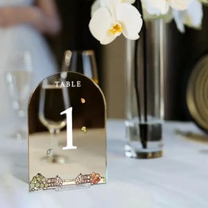 May include: A gold mirrored table number with a white number "1" and the text "TABLE" on top. The number is decorated with small gold and orange leaves. The table number is on a white table.