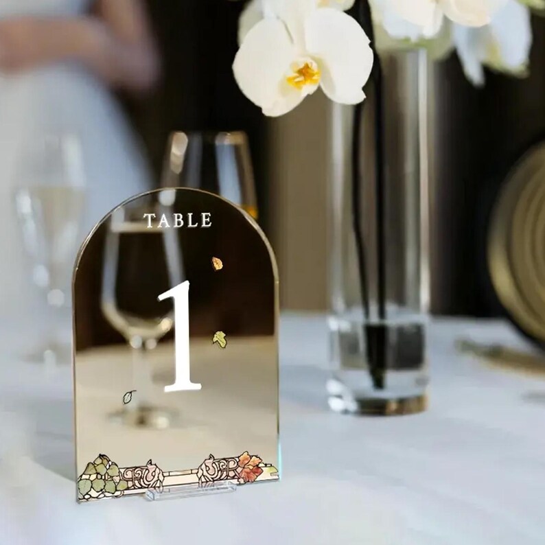 May include: A gold mirrored table number with a white number "1" and the text "TABLE" on top. The number is decorated with small gold and orange leaves. The table number is on a white table.