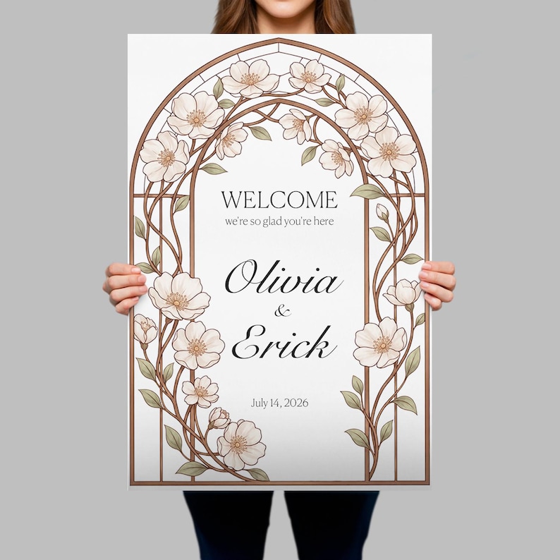 K&ouml;nnte beinhalten: Ein rechteckiges Hochzeits-Willkommensschild mit braunem Bogenrahmen, wei&szlig;en Bl&uuml;ten und gr&uuml;nen Bl&auml;ttern. Der Text lautet: WELCOME we're so glad you're here, Olivia & Erick, sowie das Datum 14. Juli 2026 in schwarzer Schriftart.
