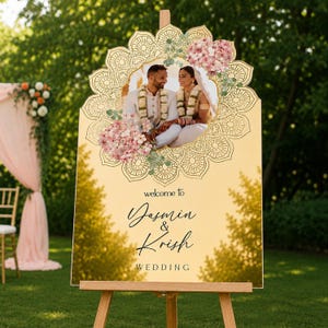 Peut inclure: Un panneau de bienvenue de mariage dor&eacute; avec une photo centrale d'un couple en tenue traditionnelle, entour&eacute; d'un motif mandala d&eacute;coratif et de fleurs roses. Le texte indique : welcome to Jasmin & Krish WEDDING.
