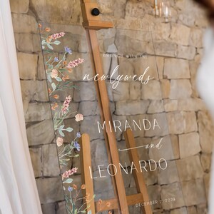 K&ouml;nnte beinhalten: Ein klares Acryl-Hochzeitschild mit floralem Design und dem Text "Brunch mit den Frischverm&auml;hlten Miranda und Leonardo 2. Dezember 2024".
