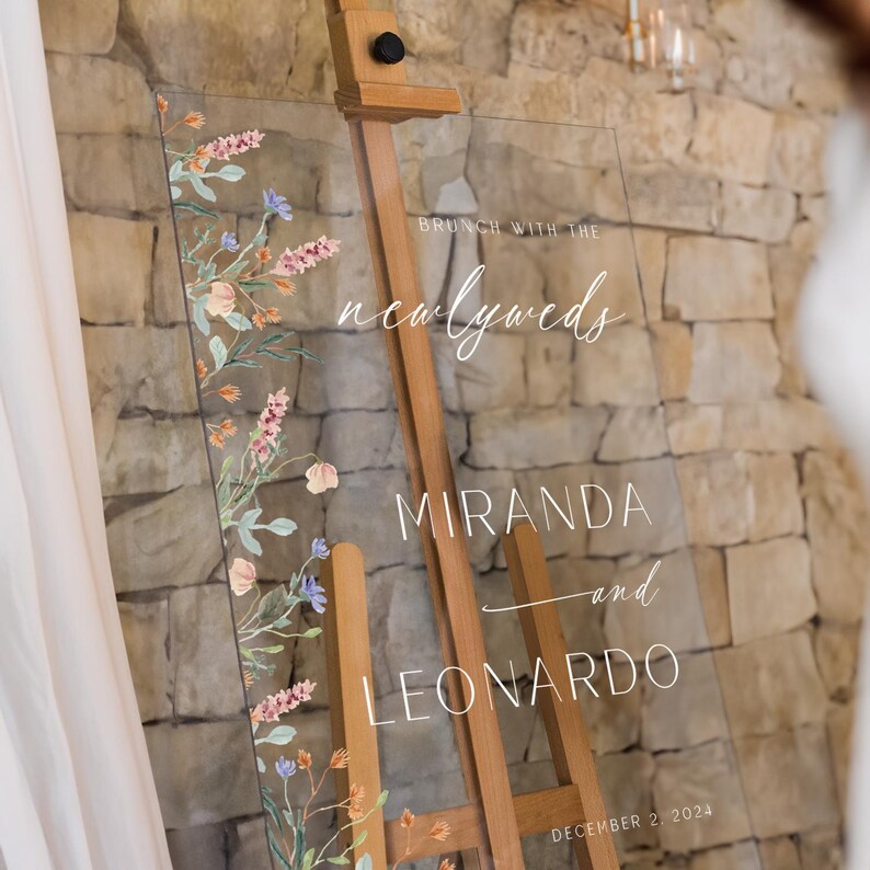 K&ouml;nnte beinhalten: Ein klares Acryl-Hochzeitschild mit floralem Design und dem Text "Brunch mit den Frischverm&auml;hlten Miranda und Leonardo 2. Dezember 2024".