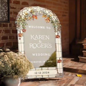 May include: A stained glass wedding welcome sign with a floral design in shades of green, orange, and brown. The sign reads "Welcome to Karen & Roger's Wedding Forever in Love 12 12 2024".