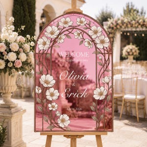 K&ouml;nnte beinhalten: Ein bogenf&ouml;rmiges Hochzeits-Willkommensschild mit rosa Hintergrund und einem floralen Rand im Buntglas-Stil. Der Text lautet: WELCOME we're so glad you're here, Olivia & Erick, July 14, 2026. Das Schild steht auf einem h&ouml;lzernen Staffeleist&auml;nder.