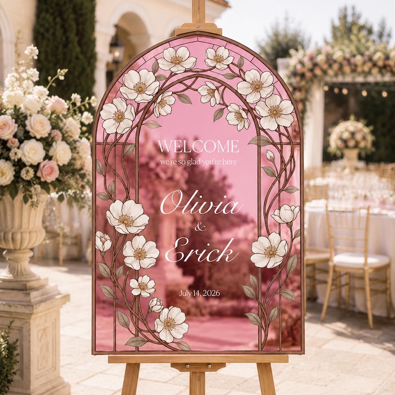K&ouml;nnte beinhalten: Ein bogenf&ouml;rmiges Hochzeits-Willkommensschild mit rosa Hintergrund und einem floralen Rand im Buntglas-Stil. Der Text lautet: WELCOME we're so glad you're here, Olivia & Erick, July 14, 2026. Das Schild steht auf einem h&ouml;lzernen Staffeleist&auml;nder.
