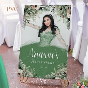 May include: A green and white floral backdrop with a photo of a young woman in a green dress. The text "Arianni's Quinceanera 11.06.24" is printed on the backdrop.