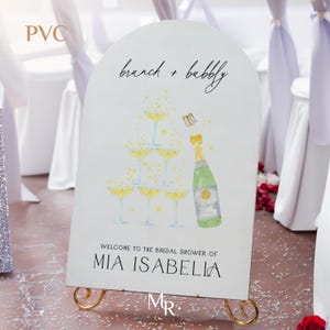 May include: A white sign with a champagne tower and bottle illustration. The sign reads "brunch + bubbly" and "Welcome to the bridal shower of MIA ISABELLA".