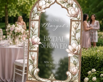 May include: An arched sign on a wooden easel featuring a stained glass border with pink magnolia flowers and green vines. The center text reads: WELCOME TO Margot's BRIDAL SHOWER NEW YORK, USA 07.18.2026. The sign has a reflective surface finish.