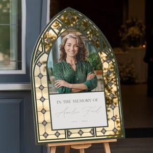 May include: A memorial sign featuring a photo of a woman in a green top, framed by a decorative arch. The sign includes the text "IN THE MEMORY OF Amelia Ford" and dates 1973-2024. The sign is displayed on a wooden easel.