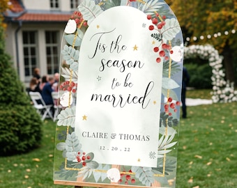 May include: An arched clear acrylic wedding sign featuring a festive botanical border of green leaves, red berries, and gold accents. The text reads 'Tis the season to be married', 'CLAIRE & THOMAS', and '12.20.22' in black script and serif fonts.