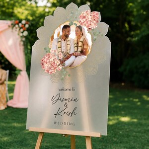 Peut inclure: Un panneau de bienvenue de mariage en acrylique d&eacute;poli avec une photo centrale d'un couple en tenue traditionnelle, orn&eacute; d'illustrations d'hortensias roses et de motifs mandala dor&eacute;s. Le texte indique, welcome to Yasmin & Krish WEDDING.